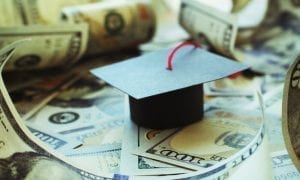 Small black graduation cap with red tassel atop a pile of curled up 100 dollar bills