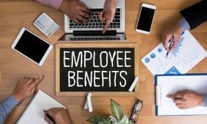 Three people's hands on desk writing on papers and using laptop with sign that says "Employee Benefits"