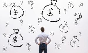 Businessman in work attire facing away from the camera looking at a wall of question marks and money bags